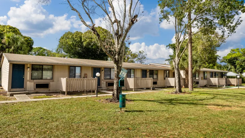 Row of single-story beige apartments with blue doors, a green lawn, and trees under a sunny blue sky.