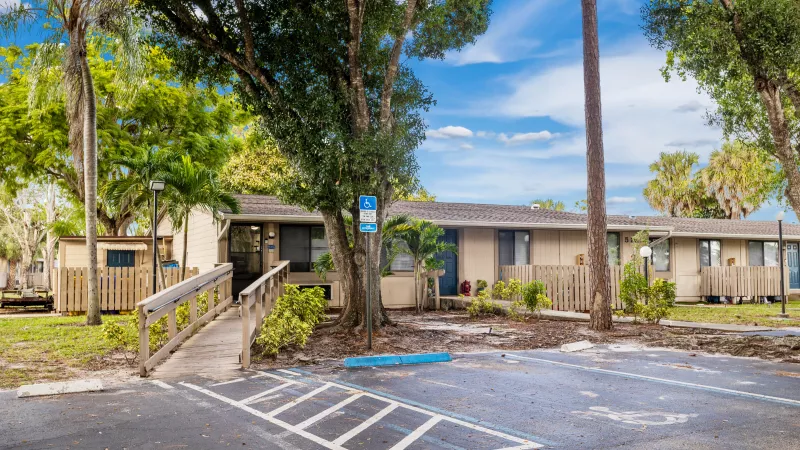 Tan residential building with a wooden access ramp and handicap parking, lush trees, blue sky.