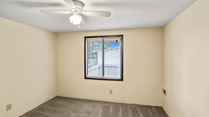Empty room with light beige walls, gray carpet, a window with blinds, and a white ceiling fan.