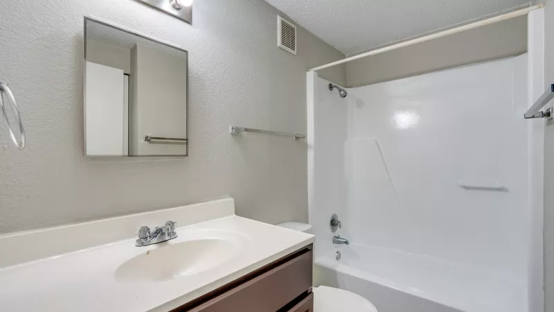 Light gray bathroom with brown vanity, white sink, mirror, toilet, and a white tub/shower combo.