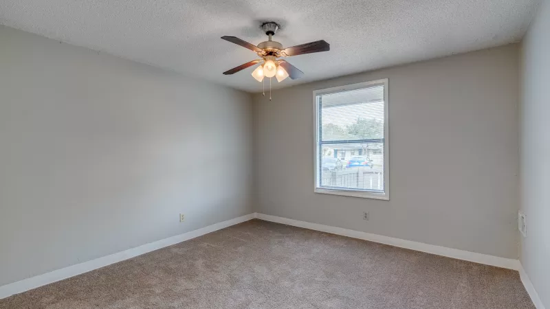 Empty bedroom with beige walls, brown carpet, a window, and a ceiling fan.