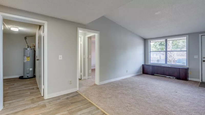 Empty living room with light gray walls, beige carpet, a window with a dark bench, and open doorways to a utility room and hallway.