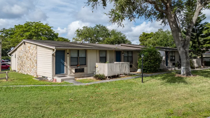 Photograph of a beige apartment building with multiple blue doors, stone accents, green grass, and trees under a sunny sky.