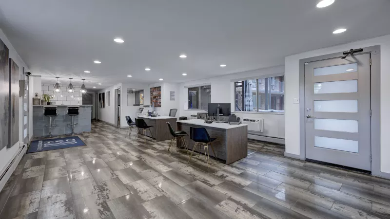 Bright, modern office interior with a kitchenette, bar seating, several computer workstations, and light gray wood-look flooring.