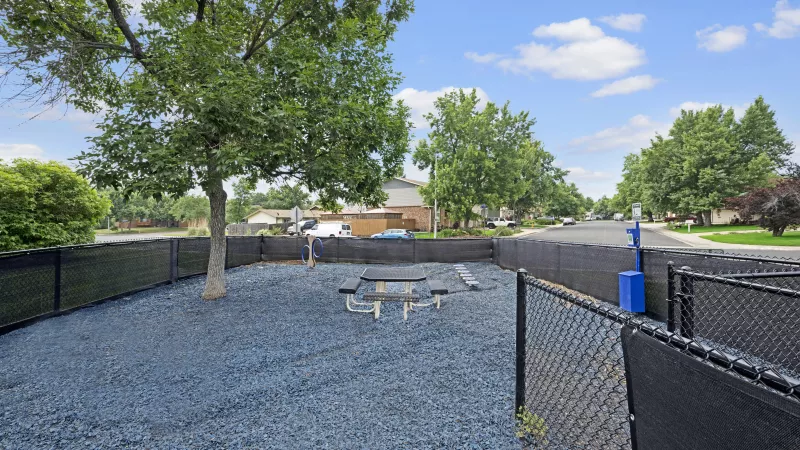 Photograph of a fenced dog run with blue gravel, a picnic table, and a tree. Residential street in the distance.