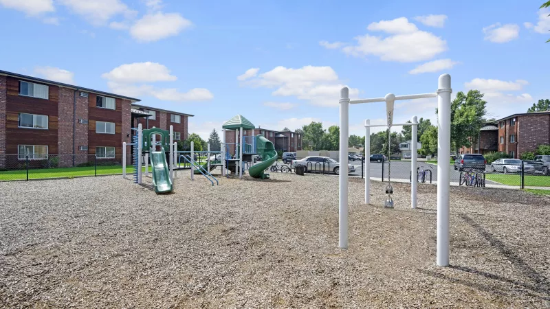 Outdoor playground with green and blue play equipment, white monkey bars, wood chips, and brick apartment buildings.