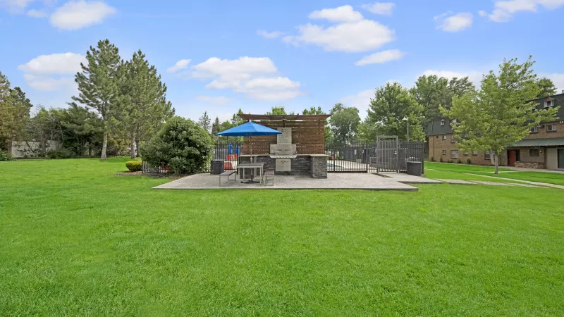 Green lawn in foreground, concrete patio with table, blue umbrella, and grill. Fenced area, trees, and buildings beyond.