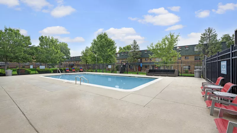 Outdoor swimming pool with red lounge chairs on a concrete deck, framed by trees and apartment buildings under a blue sky.