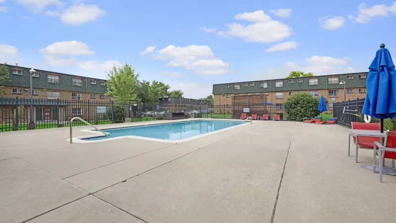 Outdoor swimming pool area with concrete deck, apartment buildings in the background, blue umbrellas, and red chairs.
