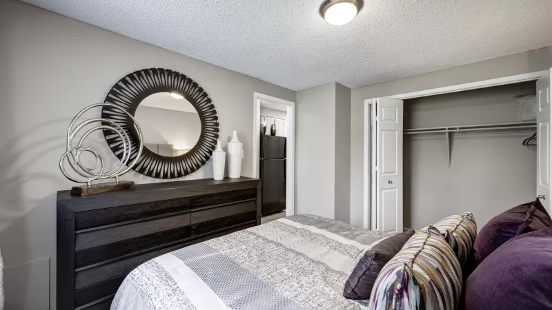 Light gray bedroom with dark wood dresser, large round ornate mirror, striped bed, purple pillows, and open closet.
