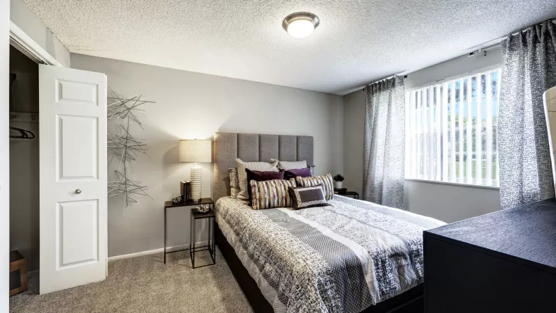 Bedroom with a queen bed, grey upholstered headboard, striped comforter, nightstand, and a window with blinds.