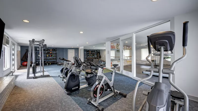 A well-lit home gym with exercise bikes, an elliptical, weight machines, and free weights.