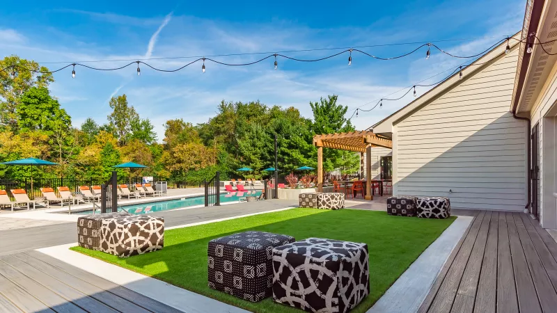 Outdoor apartment complex recreation area with pool, lounge chairs, artificial turf, patterned ottomans, and string lights.