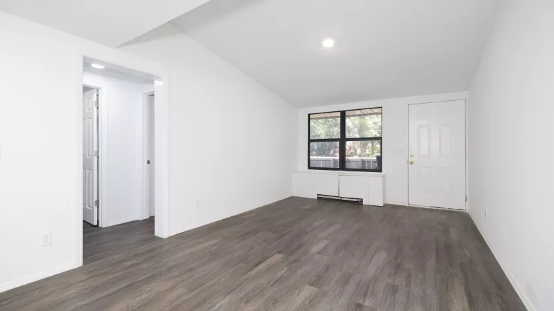 Empty room with white walls, dark laminate floor, a large window showing trees, and a white door.