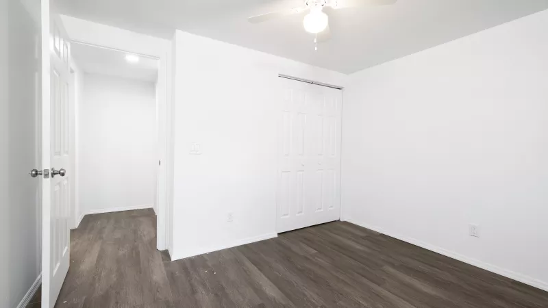 Empty room with white walls, dark wood-look floor, open doorway, closet with bifold doors, and a ceiling fan.