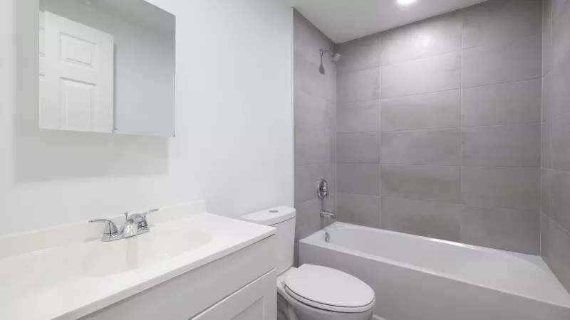 Modern bathroom with a white vanity, sink, toilet, and bathtub with gray tiled walls.