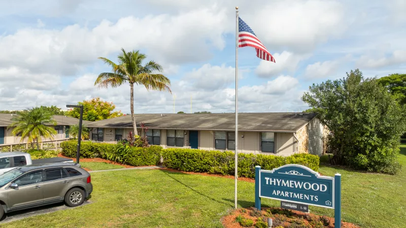 Thymewood Apartments sign with an American flag, surrounded by green lawns, palm trees, low-rise buildings, and parked cars.