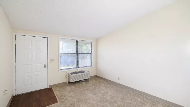 Empty beige room with a white door, window, wall-mounted AC unit, and light brown carpet.