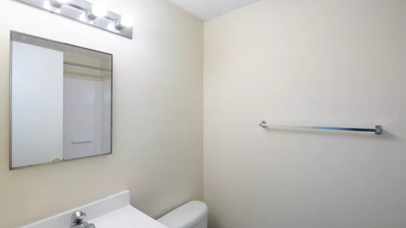 Brightly lit small bathroom with a white vanity, sink, mirror, and toilet. A towel rack hangs on the light-colored wall.