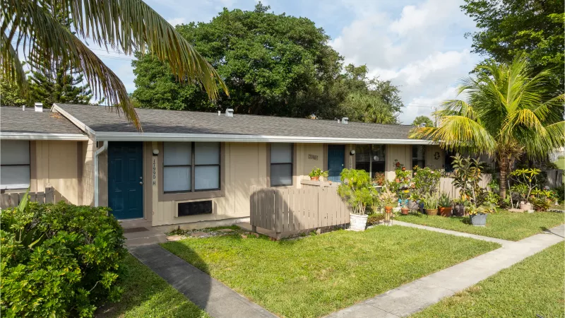 Beige single-story apartment units with teal doors, green lawns, and palm trees under a partly cloudy sky.