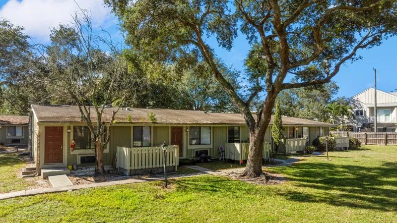 Light green, one-story apartment complex with a grassy yard and large trees under a bright blue sky.
