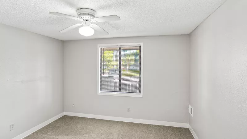 Unfurnished room with light gray walls, beige carpet, a white ceiling fan, and a window overlooking a patio.