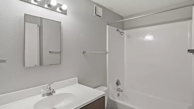 Small bathroom with light gray walls. Features a white vanity, sink, mirror, and a bathtub with shower.