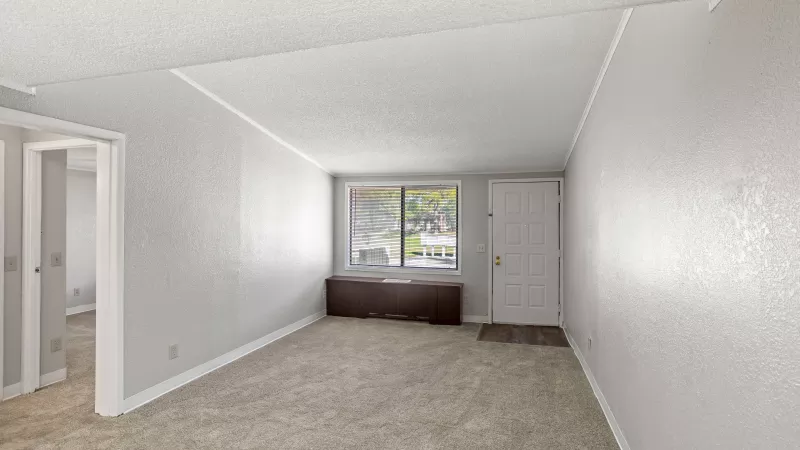 Empty living room with light gray walls, beige carpet, a large window, dark brown built-in bench, and a white entry door.