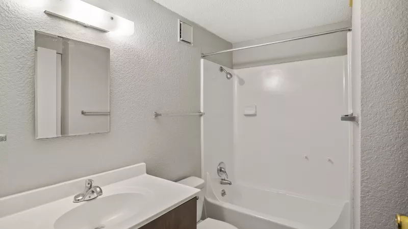 Clean, modern bathroom with a white vanity, sink, mirror, toilet, and shower-tub combo. Light grey patterned walls.