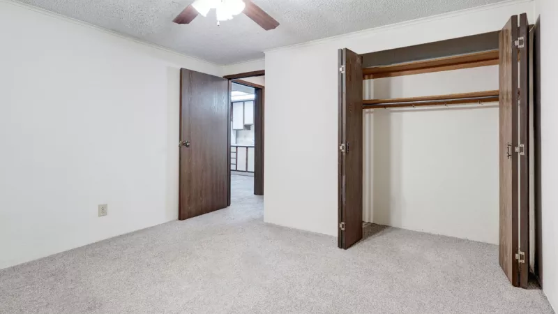 Empty bedroom with light gray carpet, white walls, an open wooden closet, and a ceiling fan.