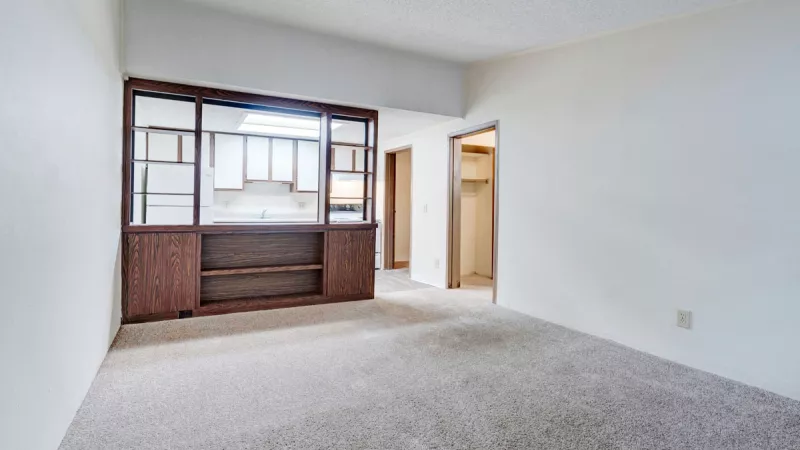 Empty living room with light carpet, white walls, and a dark wood built-in shelf looking into a kitchen.