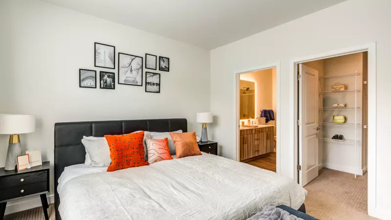 Modern bedroom with a black bed, orange pillows, and open doorways to a bathroom with vanity and a walk-in closet.