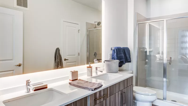 Bright modern bathroom with a double sink vanity, large back-lit mirror, and glass shower.