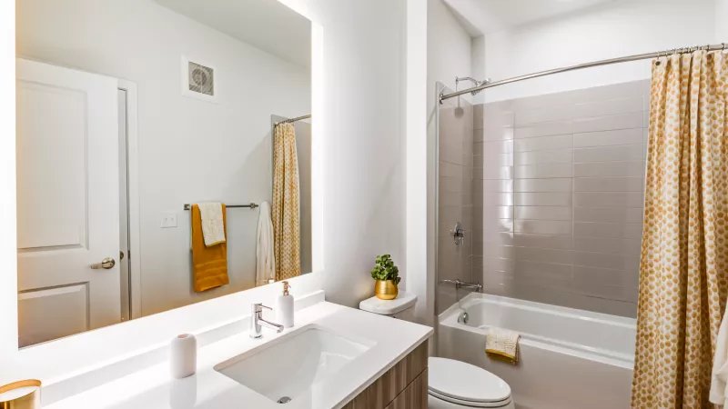 Modern bathroom with a white vanity, backlit mirror, toilet, and bathtub with a gold patterned shower curtain and towels.