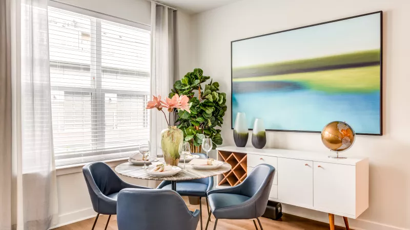 Modern dining room features a round marble table with blue chairs, a white buffet, and a large abstract painting.