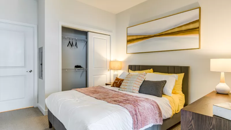 Bright bedroom featuring a gray bed with colorful pillows, a pink throw, and abstract gold art. Open closet.
