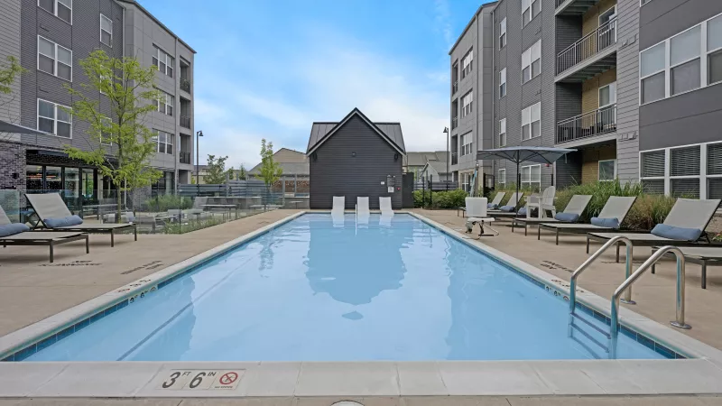 Outdoor swimming pool with lounge chairs between two modern apartment buildings and a dark gabled clubhouse under a blue sky.