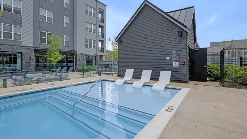 Modern apartment complex pool with a dark gray building, white lounge chairs in the water, and a large residential building.