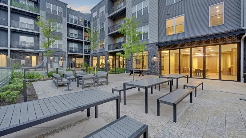 Modern apartment courtyard with grey tables, benches, lounge chairs, and small trees, surrounded by a residential building.
