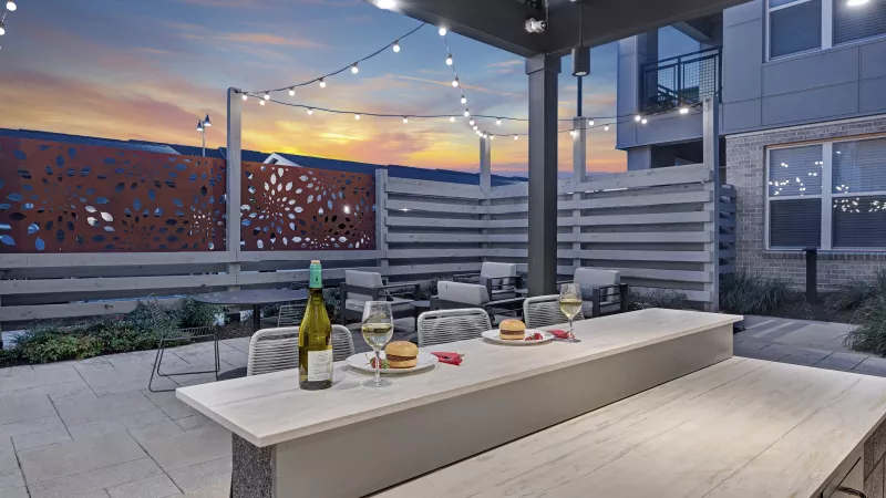 Modern outdoor patio with a long bar, burgers, wine, and string lights under a colorful sunset sky. Seating areas are visible.