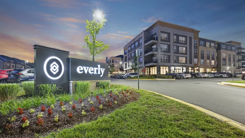 Lit Everly sign and logo in front of a modern apartment complex at dusk, with parked cars under a colorful sky.