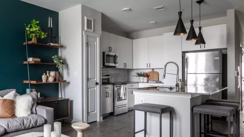 Modern open-concept living room and kitchen with a teal accent wall, floating shelves, gray sofa, white cabinets, and island.