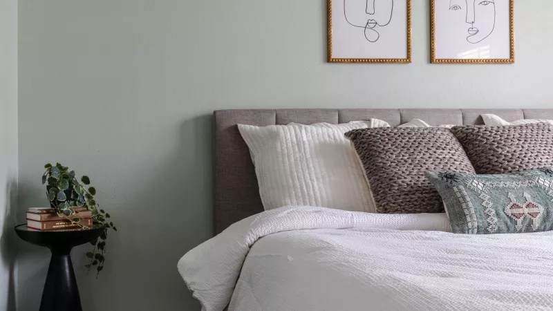 Stylish bedroom with sage green walls, a bed with white quilt and decorative pillows. Two framed minimalist face drawings hang above.