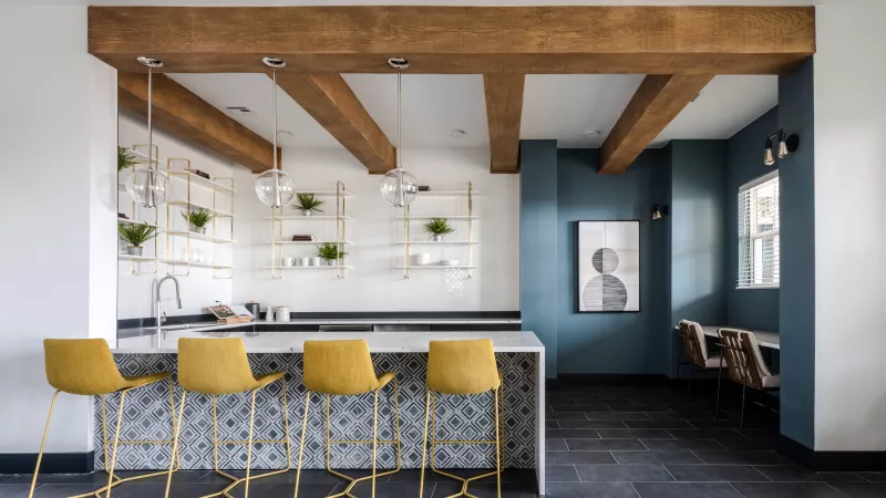 Modern kitchen features a light island with four yellow stools, wooden beams, white shelves, and a teal accent wall with artwork.