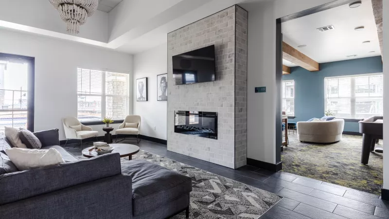 Modern living room with grey sectional, dark floor, white brick fireplace, TV, and chandelier. Opens to a blue room.