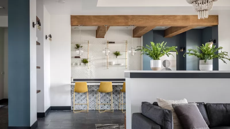 Modern open-plan living room with dark sofa, yellow bar stools, wooden beams, and a teal accent wall.