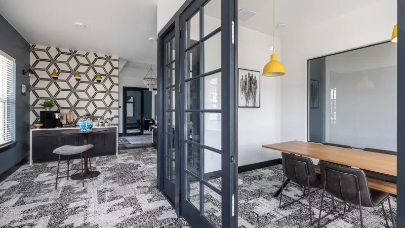 Modern office interior featuring a reception area with a geometric wall and a meeting room with a wooden table, separated by glass doors.