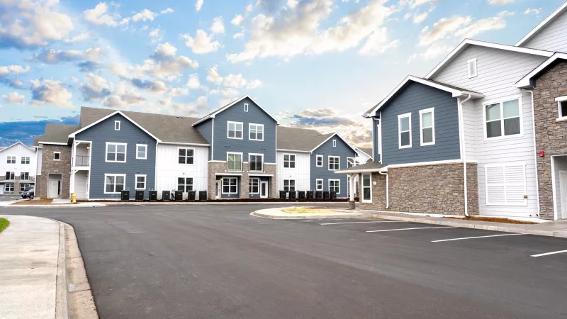 Modern residential complex with blue-gray and white buildings, a paved road, and a partly cloudy sky.