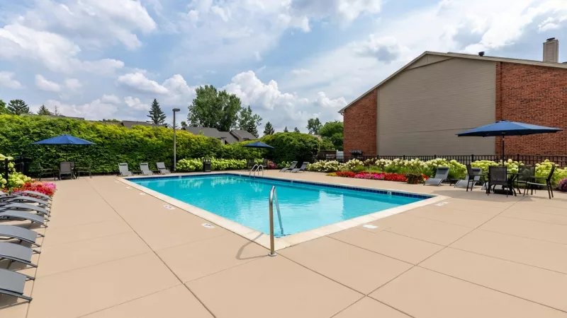 Outdoor swimming pool with blue water, lounge chairs, blue umbrellas, colorful flowers, and a brick building under a cloudy sky.