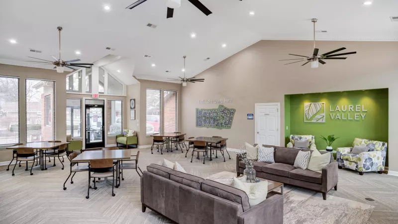 Spacious community lounge with dining tables, comfortable sofas, and a green accent wall with the Laurel Valley logo.